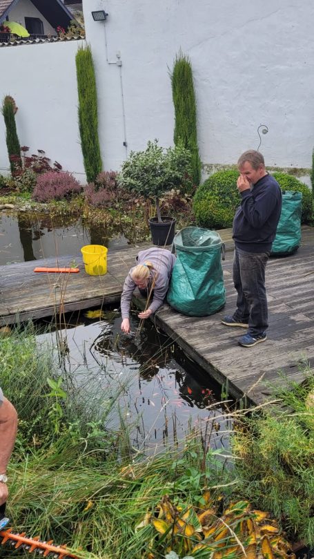 Person beugt sich über einen Teich, während eine andere daneben steht und telefoniert.