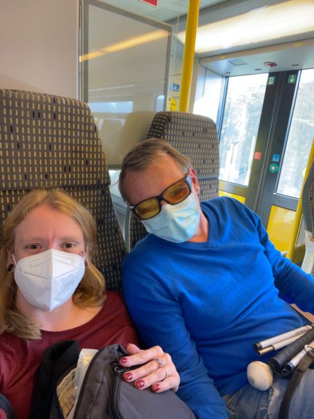 Personen mit Masken sitzen in einem Zug, eine Person trägt eine Sonnenbrille.