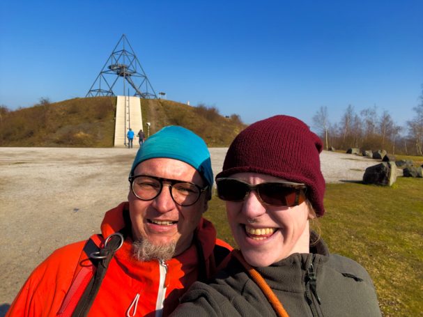 Zwei Personen lächeln vor einer Berglandschaft mit einem Stahlgerüst im Hintergrund.