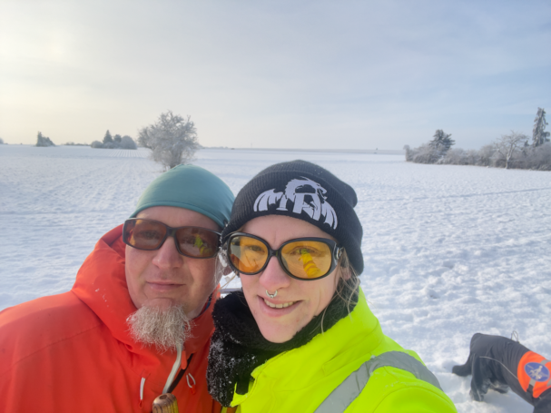 Zwei Personen in winterlicher Landschaft mit Schnee, beide tragen farbige Winterkleidung.