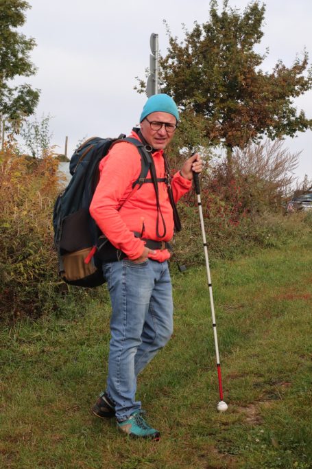 Mann in orange Jacke mit Rucksack und Gehstock, steht auf einem Wanderweg.