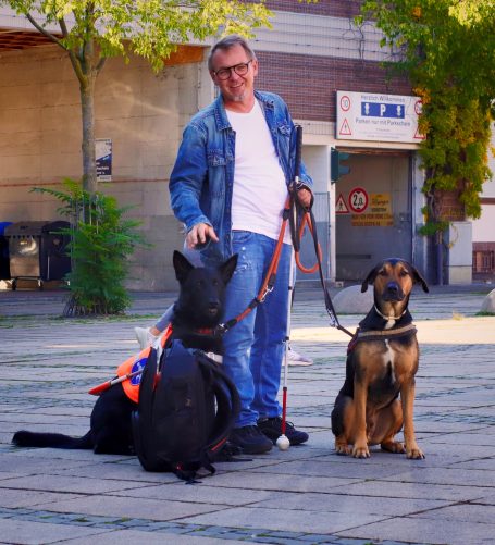 Mann mit zwei Hunden, einer davon ein Blindenführhund, in einer urbanen Umgebung.